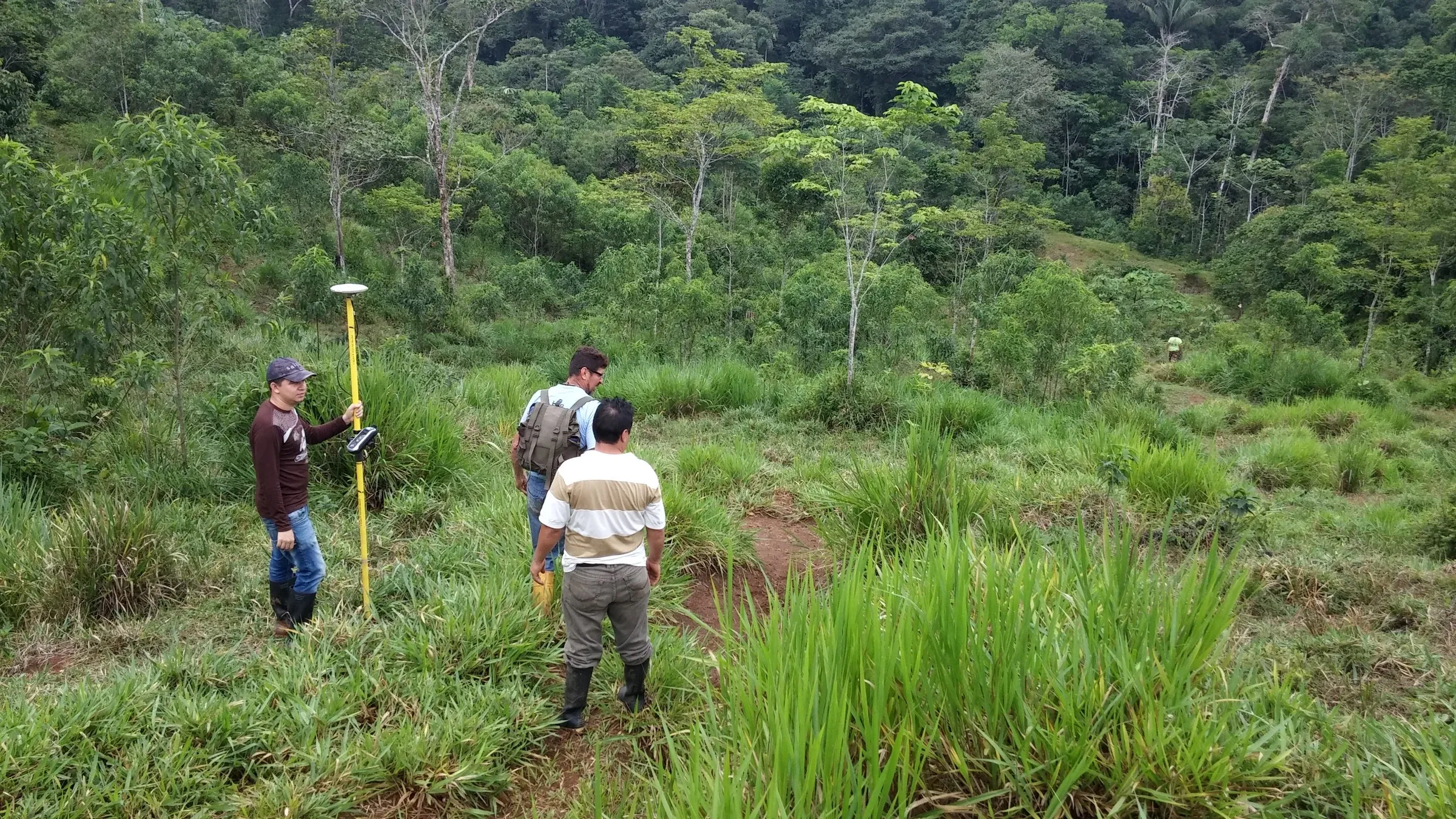 Tres personas levantan datos en un claro rodeado de selva amazónica; una sostiene un receptor GPS montado sobre un jalón.