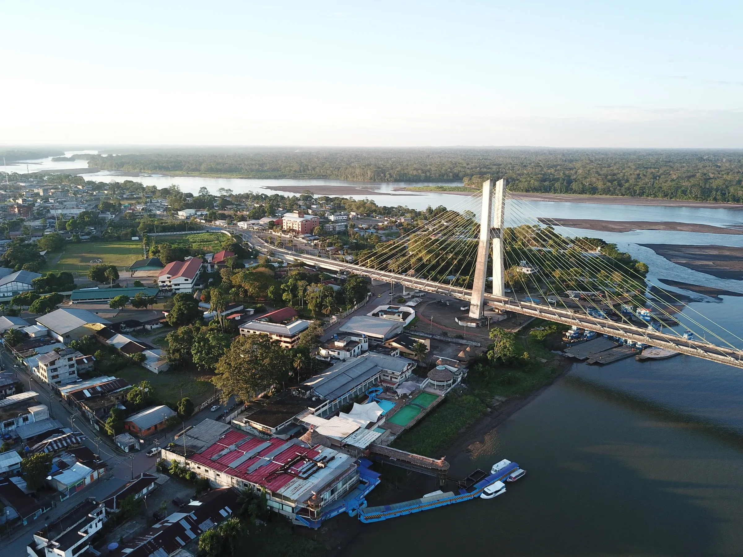 Vista aérea de El Coca al amanecer: el puente atirantado sobre el río Napo, la ciudad a un lado y la selva extendiéndose hasta el horizonte.