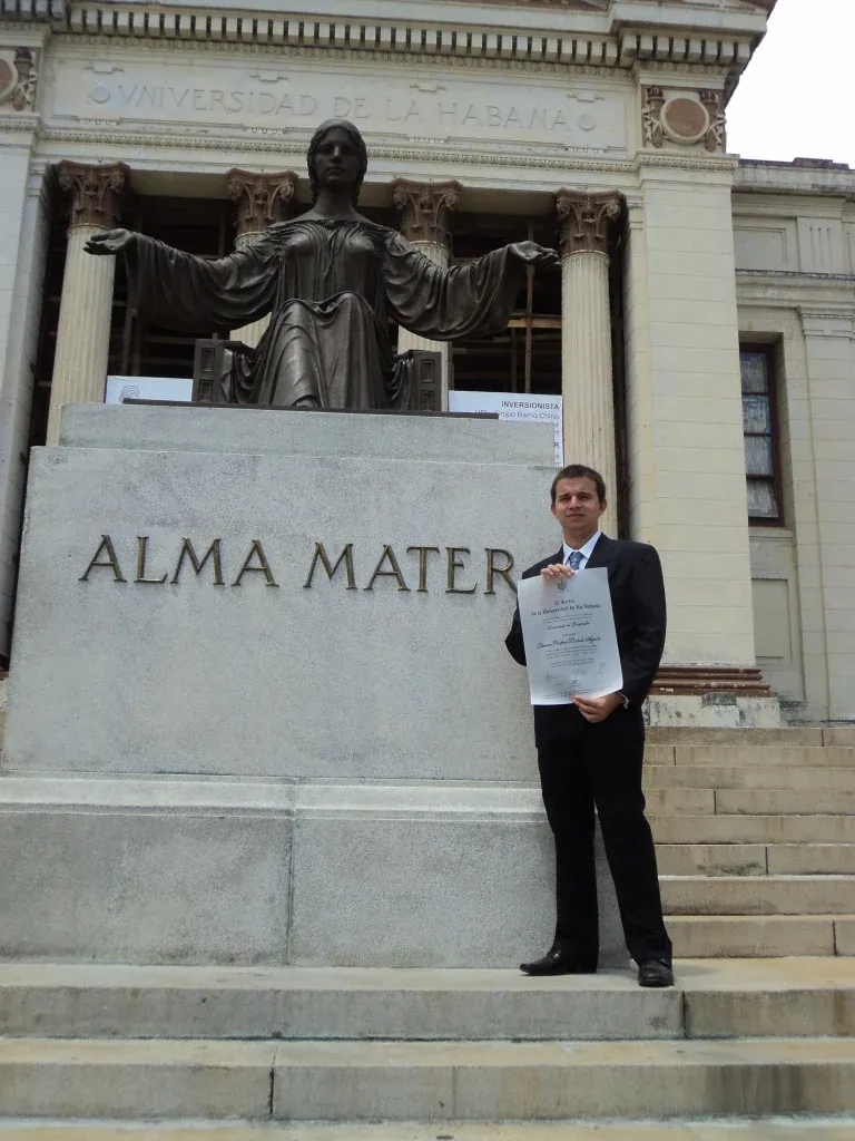 Lázaro Batule, de traje, sostiene su título de licenciatura junto a la estatua del Alma Mater, en la escalinata de la Universidad de La Habana.