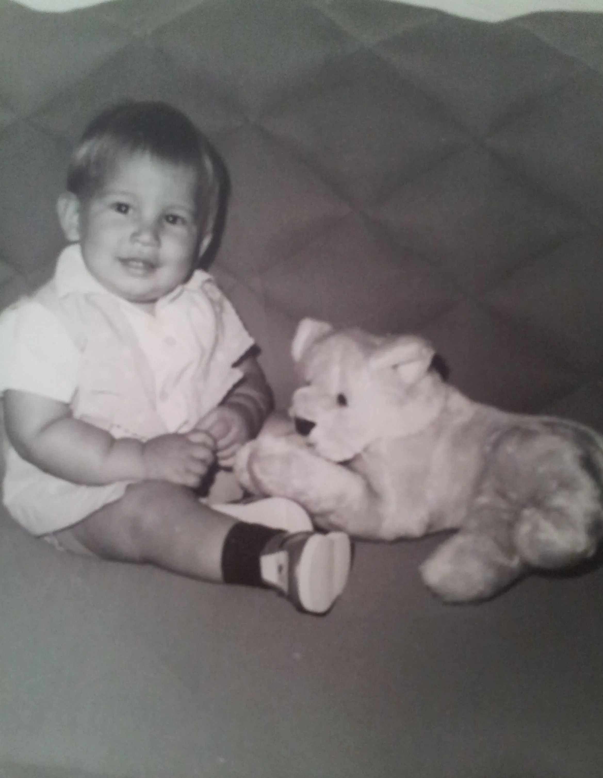 Lázaro Batule de bebé, sentado y sonriendo junto a un perro de peluche, en una fotografía en blanco y negro.