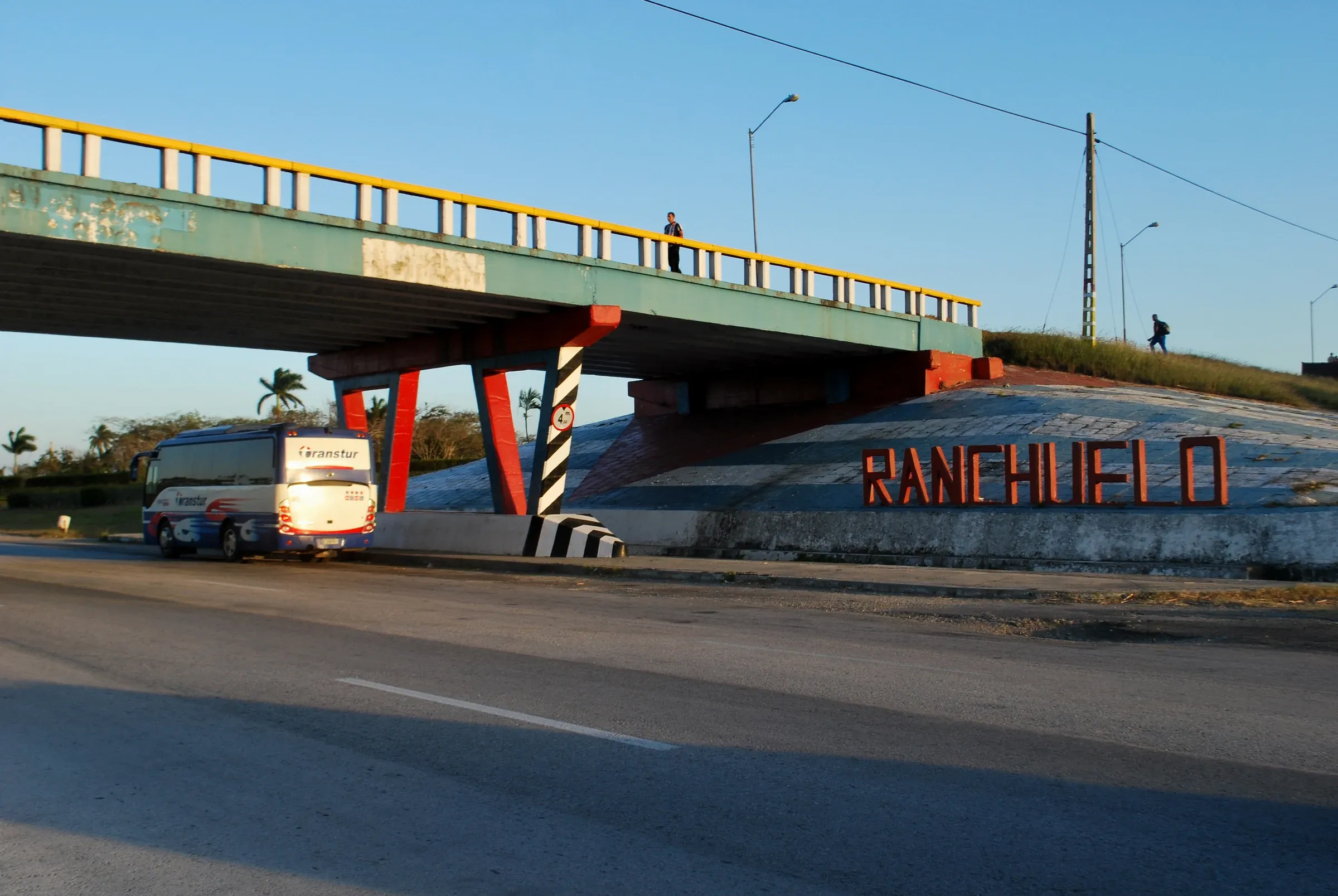 El nombre de Ranchuelo escrito en grandes letras rojas sobre el terraplén de un puente de carretera, con un ómnibus detenido al costado, a la luz del atardecer.